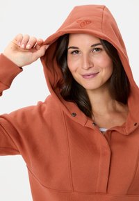 Sweat à capuche couleur rouille avec une grande capuche ajustable, présentant un tissu texturé et des boutons pression au niveau du col.