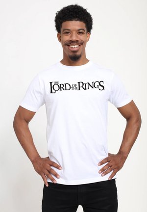 Lächelnder Mann steht mit den Händen in den Hüften, trägt ein weißes T-Shirt mit dem Aufdruck "Der Herr der Ringe" auf der Vorderseite.