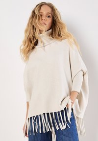 Poncho color crema con un colletto alto, realizzato in tessuto morbido, caratterizzato da frange laterali e dettagli a coste sui polsini.