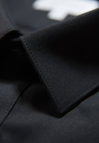 Zwarte stoffen kraag van een shirt met een textuur, voorzien van een gestikt randje en een strakke, puntige vorm. Subtiel contrast met witte accenten.
