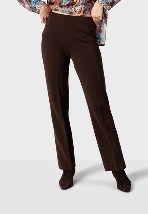 Personne portant un pantalon droit marron foncé et des bottines foncées, debout devant un fond uni.