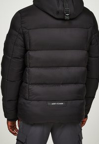Schwarze Steppjacke mit horizontalen Steppnähten, Kapuze, verstellbarem Riemen und Markenpatch auf der Rückseite. Glattes, leichtes Material.