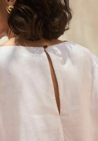 Bílá lněná blůza s otvorem na zádech ve tvaru klíče a dekorativním knoflíkem. Hladká textura s lehkými, splývavými rukávy. Jednoduchý a minimalistický design.