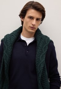 Donkerblauwe gebreide trui met kraag, met een donkergroene kabelgebreide sjaal over de schouders gedrapeerd, tegen een neutrale achtergrond.