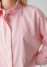 Light pink button-up shirt with a collar, relaxed fit, long sleeves, and a smooth, slightly wrinkled texture. Subtle sheen visible.
