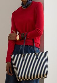 Άτομο που φοράει κόκκινο πουλόβερ και μπλε τζιν, κρατώντας μια τσάντα χειρός με ναυτικό και λευκό ριγέ σχέδιο με χρυσές λεπτομέρειες και φορώντας χοντρά βραχιόλια.