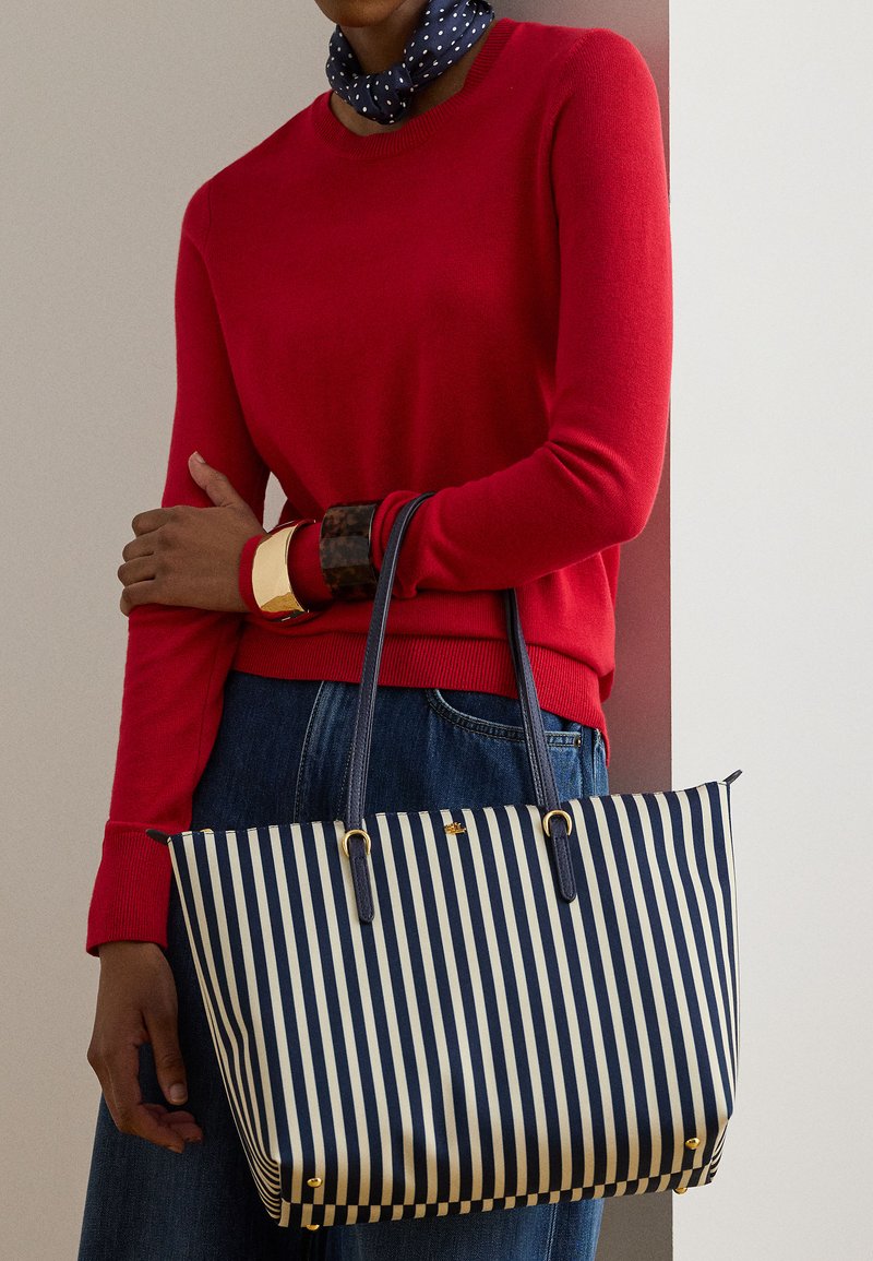 Άτομο που φοράει κόκκινο πουλόβερ και μπλε τζιν, κρατώντας μια τσάντα χειρός με ναυτικό και λευκό ριγέ σχέδιο με χρυσές λεπτομέρειες και φορώντας χοντρά βραχιόλια.