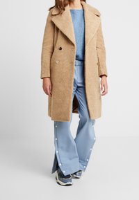 Cappotto beige texture con ampi rever e bottoni doppi, abbinato a pantaloni a gamba larga blu chiaro con dettagli in bottoni a scatto.