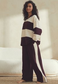 Komplet prostega domačega oblačila z ožemajočimi bordo in kremnimi črtami, ki vključuje dolg rokav s topom v obliki črke V in hlače s stransko črto. Mehka, sproščena kroj.