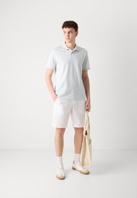 Polo shirt bleu clair à manches courtes, shorts blancs et baskets beiges. Le modèle tient un sac fourre-tout beige, avec une texture de tissu lisse.