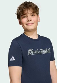 Námorníckomodrá bavlnená tričká s krátkymi rukávmi, na ktorých je vyšitý nápis "Real Madrid" svetlým písmom a biele logo Adidas na ľavom rukáve.
