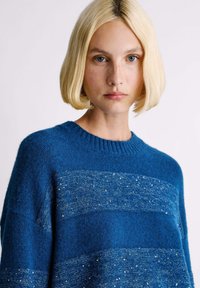 Suéter azul de punto con un acabado texturizado, presenta franjas metálicas horizontales y un cuello redondo acanalado. Material suave y cómodo.
