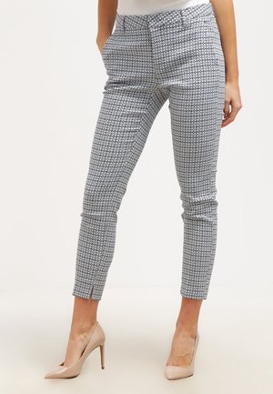 Žena nosící úzké kotníkové kalhoty s geometrickým černo-bílým vzorem a tělové vysoké podpatky, ruce v kapsách.