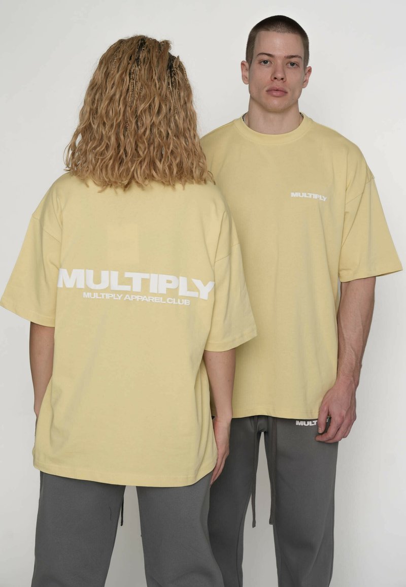 Gelbe übergroße T-Shirts mit dem weißen Aufdruck "MULTIPLY" auf der Rückseite. Kombiniert mit grauen Jogginghosen. Weiches Baumwollmaterial, lockere Passform.