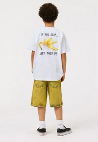 Vit t-shirt med en gul banan grafik och text på baksidan. Bärs med gula denimshorts och svarta skor med vita strumpor.