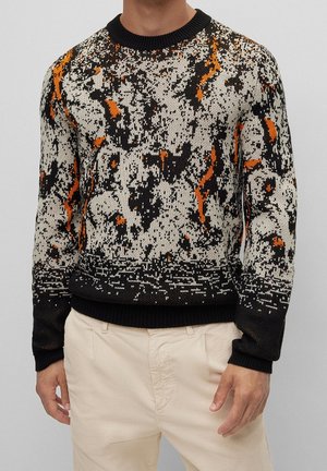 Mann trägt einen langärmligen Rundhalspullover mit einem abstrakten Muster in Schwarz, Weiß und Orange, kombiniert mit hellbeigen Hosen.