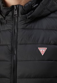 Črna puffer jakna z visokim ovratnikom, ki ima sprednji zadrgo in vodoravno quilted zasnovo. Logotip Guess je jasno izpostavljen.