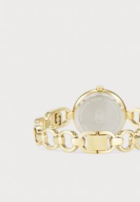 Gold watch with a linked chain bracelet, round face, and a transparent back displaying internals. Features polished metal surface and clasp.