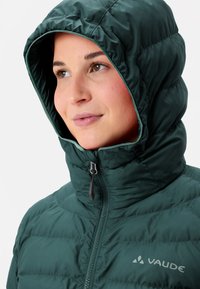 Junge Frau in einer dunkelgrünen, gepolsterten Kapuzenjacke, die bis zum Hals zugezippt ist, mit neutralem Gesichtsausdruck, leicht nach rechts schaut.