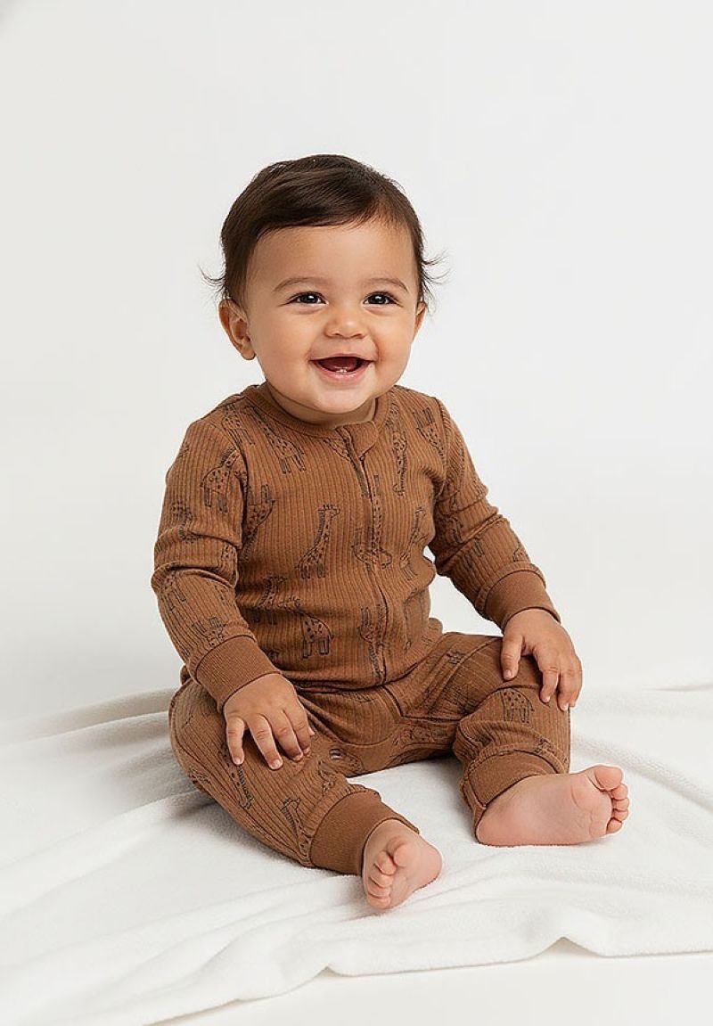 Bébé souriant aux cheveux foncés assis sur une couverture blanche, portant une grenouillère marron à manches longues avec un motif girafe, pieds nus.
