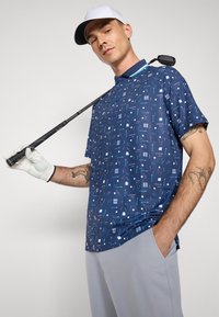 Közel félhosszú ujjú sötétkék golf póló, világoskék geometrikus mintával, szürke nadrággal és fehér sapkával kombinálva, golfütőt tartva.
