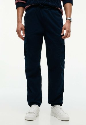 Mann trägt marineblaue Cargohosen, weiße Sneakers, einen dunklen Pullover mit roten und weißen Bündchen und eine silberne Armbanduhr am linken Handgelenk.