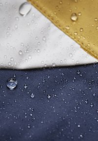 Textilie s voděodolným povrchem, obsahující béžové, bílé a námořnické sekce. Na látce jsou viditelné kapky vody, které zdůrazňují strukturu.