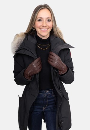 Schwarzer Parka mit pelzgefütterter Kapuze, darüber ein schwarzer Rollkragenpullover getragen. Braune Lederhandschuhe mit detaillierten Nähten. Dunkle Jeans aus Denim vervollständigen den Look.