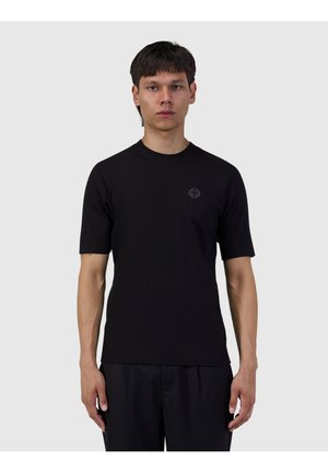 Jonge man met kort donker haar, die een effen zwart T-shirt met korte mouwen en een zwarte broek draagt, staande tegen een lichtgrijze achtergrond.