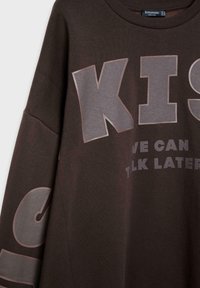 Sweat-shirt marron foncé avec de grandes lettres grises « KIS » sur le devant et un texte partiel « WE CAN TALK LATER », avec également des lettres grises sur la manche gauche.