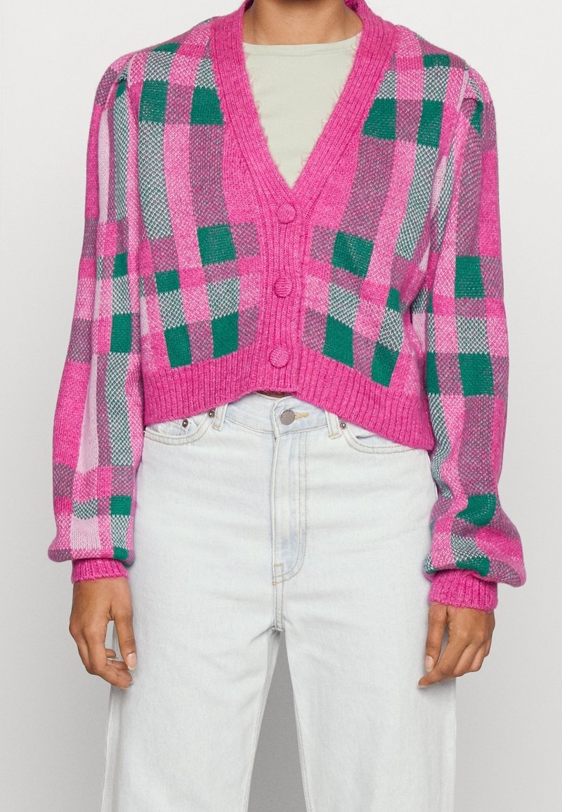 Personne portant un cardigan à carreaux rose, vert et gris avec de gros boutons sur une chemise claire et un jean taille haute bleu clair.