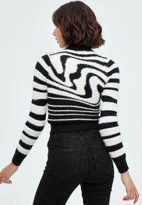 Suéter de rayas blanco y negro con una textura suave; presenta patrones ondulados y un dobladillo acanalado. Se lleva con jeans negros de cintura alta.