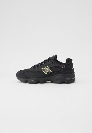 Czarny sneaker New Balance z grubą podeszwą, beżowe logo po boku, sznurowany fason i metka z materiału Cordura blisko pięty na jednolitym tle.