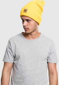 Căciulă tricotată galbenă cu o manșetă pliată și un patch dreptunghiular maro. Purtați-o cu un tricou cu mâneci scurte gri.