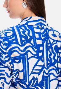 Weißes Hemd mit lebhaftem blauen abstrakten Muster, mit einem Kragen-Design und einem silbernen Knopfdetail auf der Rückseite. Strukturierter Stoff verleiht Tiefe.