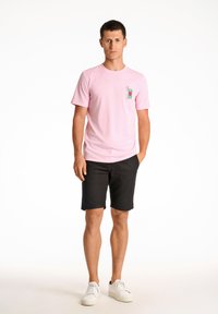 Ljusrosa t-shirt i bomull med grafisk fickdesign, matchad med svarta shorts och vita sneakers. Modellen står framför en neutral bakgrund.