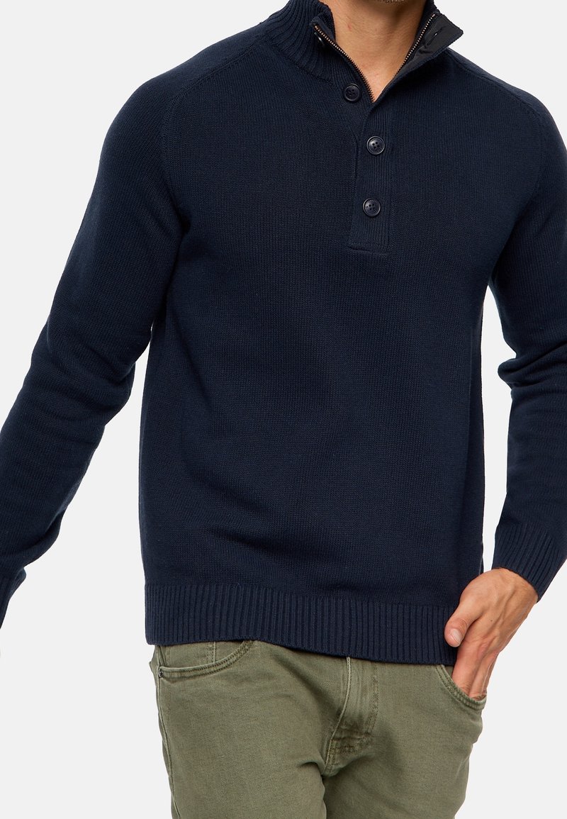 Homme portant un pull à manches longues bleu marine avec un col côtelé et trois boutons, associé à un pantalon vert olive.