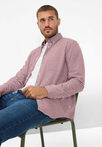 Kariertes rot-weißes Langarmhemd mit Button-Down-Kragen, kombiniert mit blauer Jeans, sitzend auf einem grünen Stuhl, schlichter Hintergrund.