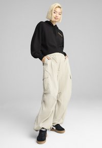 Felpa nera con logo dorato, abbinata a pantaloni cargo beige larghi e sneaker neri. Texture morbida, silhouette rilassata, tasche sui pantaloni.