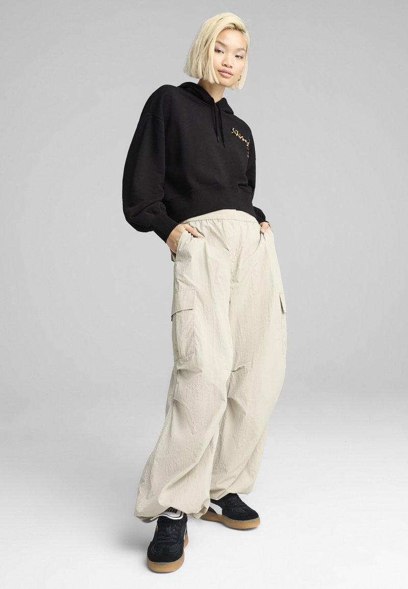 Felpa nera con logo dorato, abbinata a pantaloni cargo beige larghi e sneaker neri. Texture morbida, silhouette rilassata, tasche sui pantaloni.