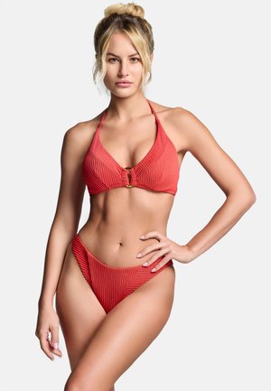 Kobieta o blond włosach w czerwonym prążkowanym bikini z przednim zapięciem, pozująca z jedną ręką na biodrze na jasnym jednolitym tle.