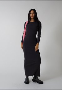 Grå figursydd klänning med långa ärmar i mjukt tyg, kombinerad med svarta texturerade ankellånga stövlar och en rosa axelväska. Enfärgad bakgrund.