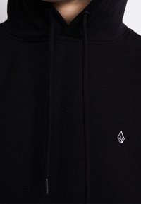 Zwarte hoodie van zacht materiaal met een trekkoordsluiting. Heeft een klein wit logo op de linker borst.
