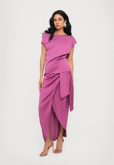 Robe fuchsia avec tissu drapé, décolleté asymétrique et détail de noeud sur le côté. Textura lisse, longueur de sol, associée à des sandales à talons nus.