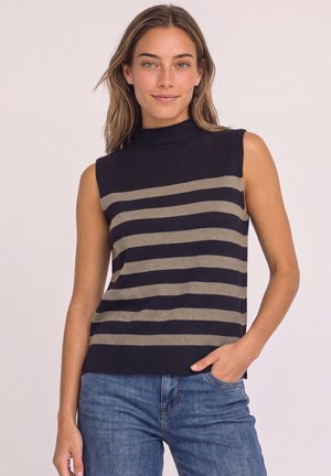 Femme portant un pull sans manches rayé noir et beige à col montant et un jean bleu, debout avec une main dans la poche sur fond uni.