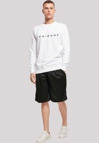 Felpa bianca con logo "FRIENDS" in nero, abbinata a pantaloni sportivi neri. Il modello indossa sneaker bianche. Tessuto liscio, vestibilità comoda.