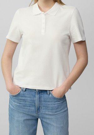 Person mit einfachem weißen kurzärmeligen Poloshirt und blauer Jeans mit den Händen in den Taschen, gezeigt von den Schultern bis zu den oberen Oberschenkeln.