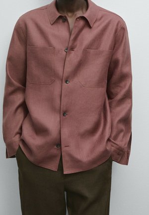 Homme portant une chemise à manches longues, boutonnée, rose poudré, avec deux poches poitrine et un pantalon foncé, debout devant un fond clair uni.