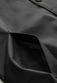Die schwarze Stoffjacke verfügt über ein weiches Innenfutter und hat eine große, schräg gelegene Tasche. Das Design ist glatt mit minimalistischem Nähen und ohne Muster.