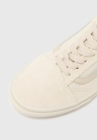 Beige suede sneaker with a rounded toe, flat laces, and a side stripe detail in a lighter shade. Features textured fabric and rubber sole.